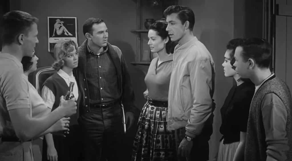A group of six young people in 1950s attire, gathered in a room with a tense atmosphere, while one person holds a beverage.