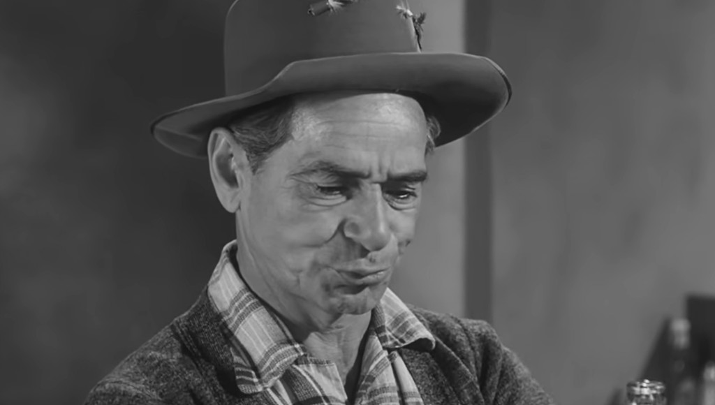 A man with a serious expression wearing a hat and a plaid shirt, looking down thoughtfully.
