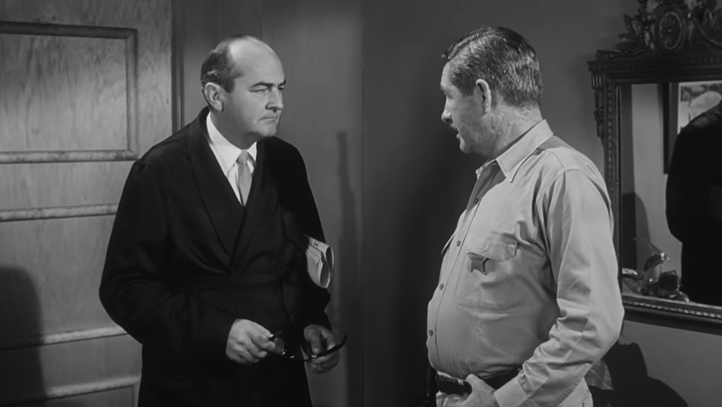 Two men in a dialogue in a room, one wearing a dark suit and holding glasses, the other in a light-colored shirt.