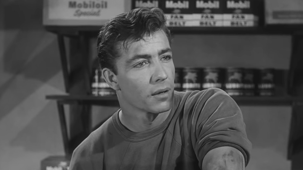 A black and white portrait of a man with tousled hair and a contemplative expression, sitting in a store with shelves of products in the background.