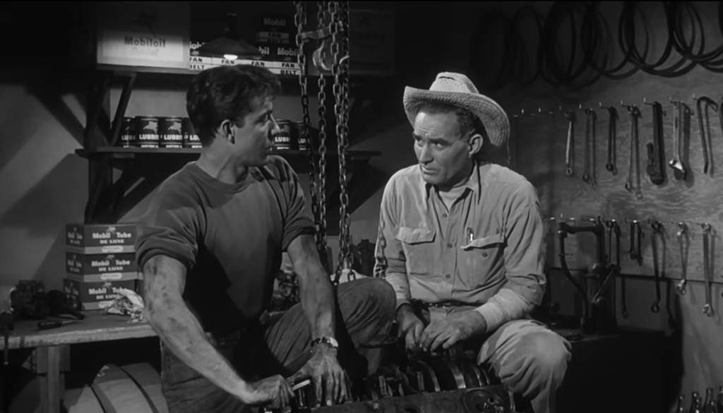 Two men engaged in conversation in a workshop, one has oil-stained hands and is seated next to a mechanical engine part, the other is wearing a cowboy hat and leaning forward.