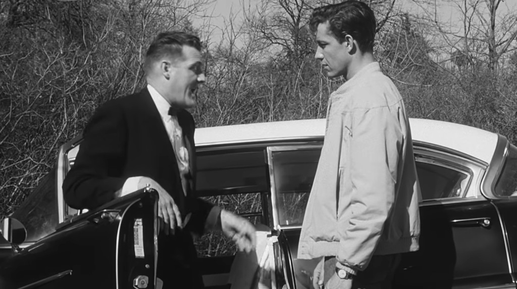 Two men stand near a vintage car in a black and white scene, one man in a suit appears to be speaking animatedly, while the other, dressed casually, looks on with a serious expression.