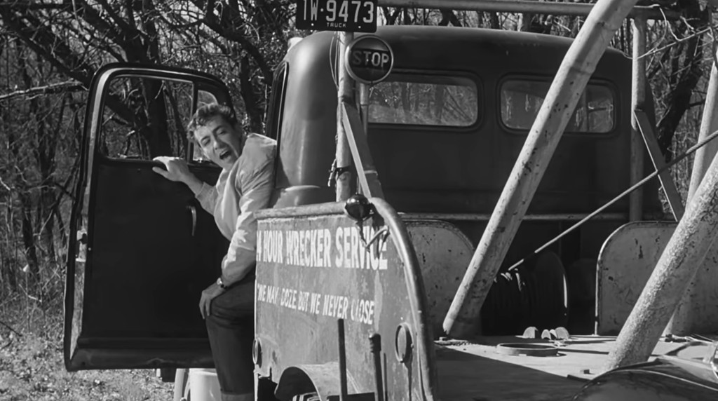 A man sitting on the side of a vintage tow truck, looking surprised or concerned, with trees in the background.