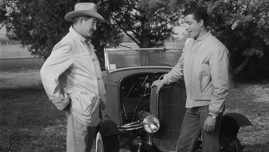 Two men are standing beside a vintage car in a rural setting, engaging in conversation. One man is wearing a light-colored jacket and a cowboy hat, while the other is dressed in a bomber jacket and jeans.