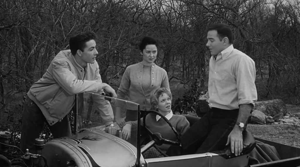 A group of four people gathered around a vintage car in a wooded area, discussing something with expressions of curiosity and concern.