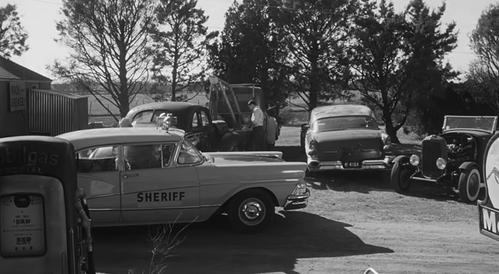 A vintage sheriff's car parked in a rural area with several classic cars, trees, and a gas station visible in the background.