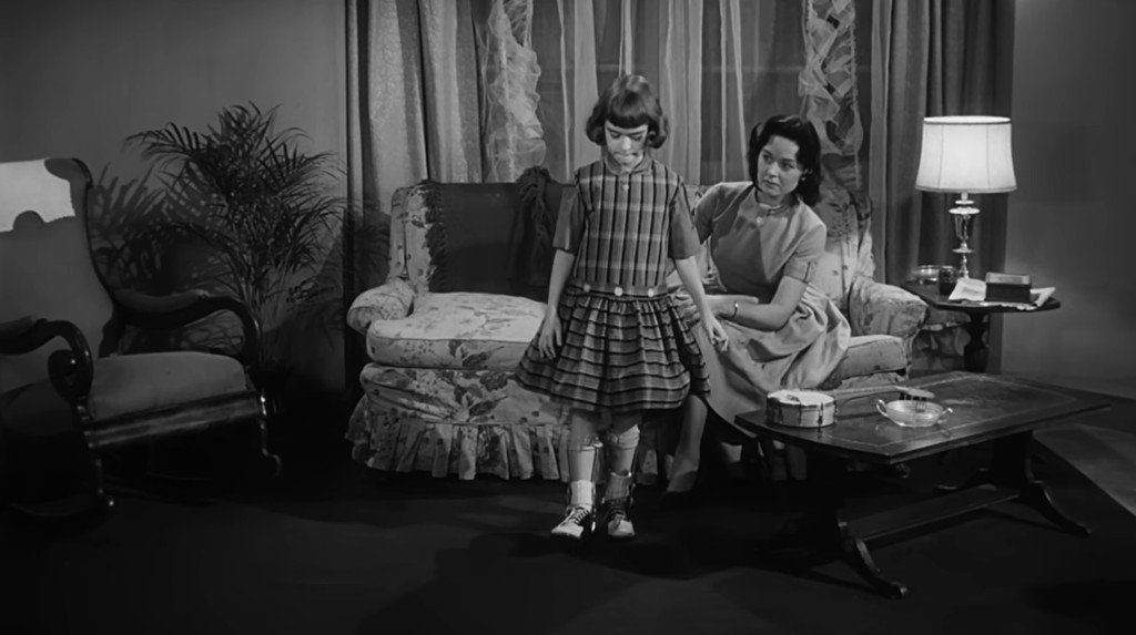 A young girl in a striped dress stands in a living room, looking down at her shoes, while an adult woman sits next to her, appearing supportive.
