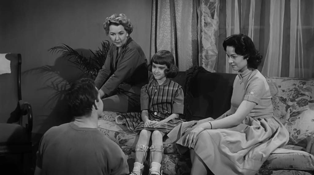 A group of four people sitting together in a living room, with three women and a young girl on a couch. The girl is wearing a plaid dress and leg braces, while the women are dressed in vintage clothing and observing a man who is seated on the floor facing them.