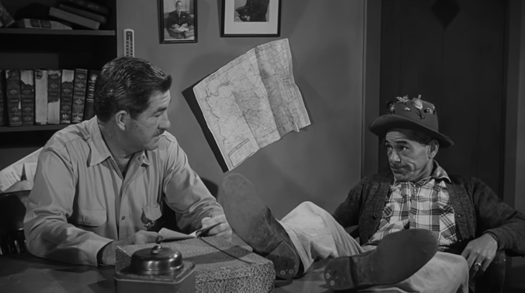 Two men sitting at a table in an office, one in a uniform and the other wearing a hat, engaged in conversation with a map on the wall behind them.