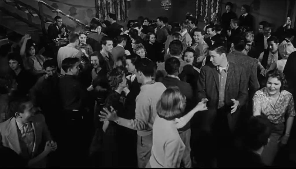 A lively dance scene in black and white, featuring a large crowd of young people enjoying themselves. The atmosphere is festive, with couples and groups dancing, smiling, and interacting in a social setting, while a DJ is seen in the background.