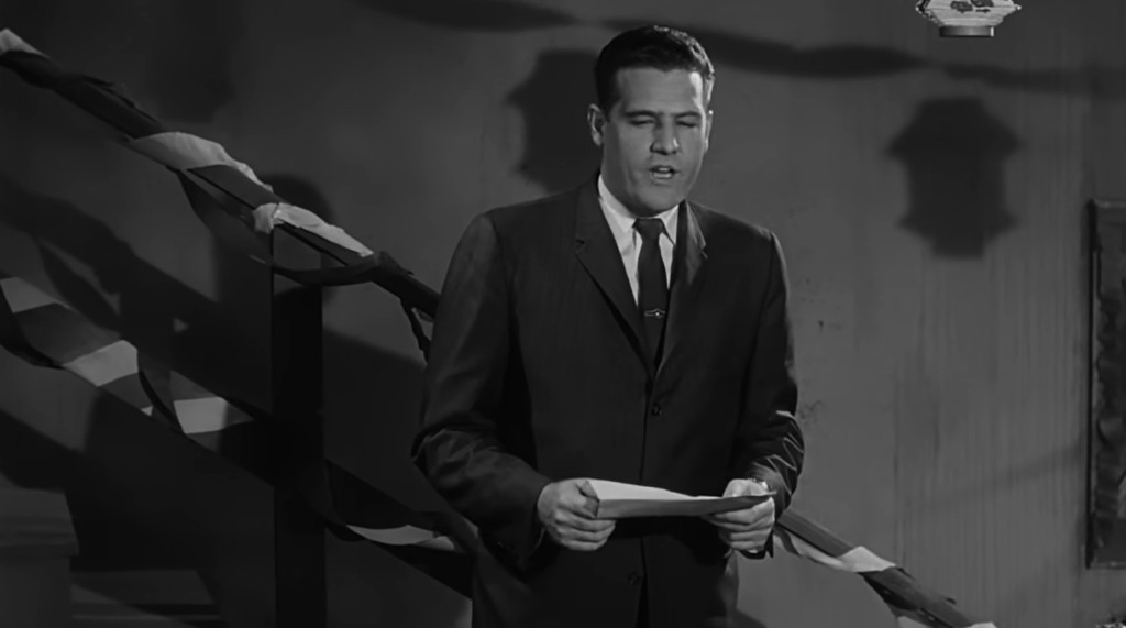 A man in a suit stands on a staircase, reading from a piece of paper, with decorations visible in the background.