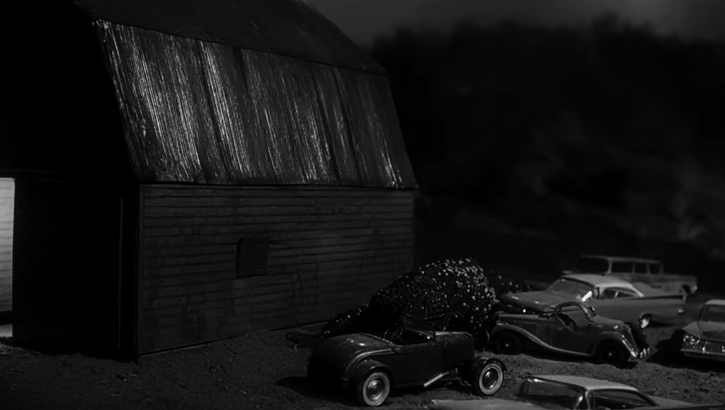A black and white image of a rustic barn with a metallic roof, surrounded by classic cars parked in a gravel area, creating a nostalgic scene.
