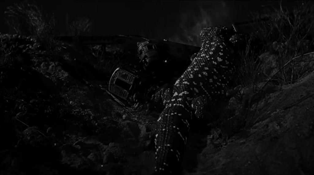 A black and white image of a lizard-like creature crawling on rocky terrain beside a mechanical object.