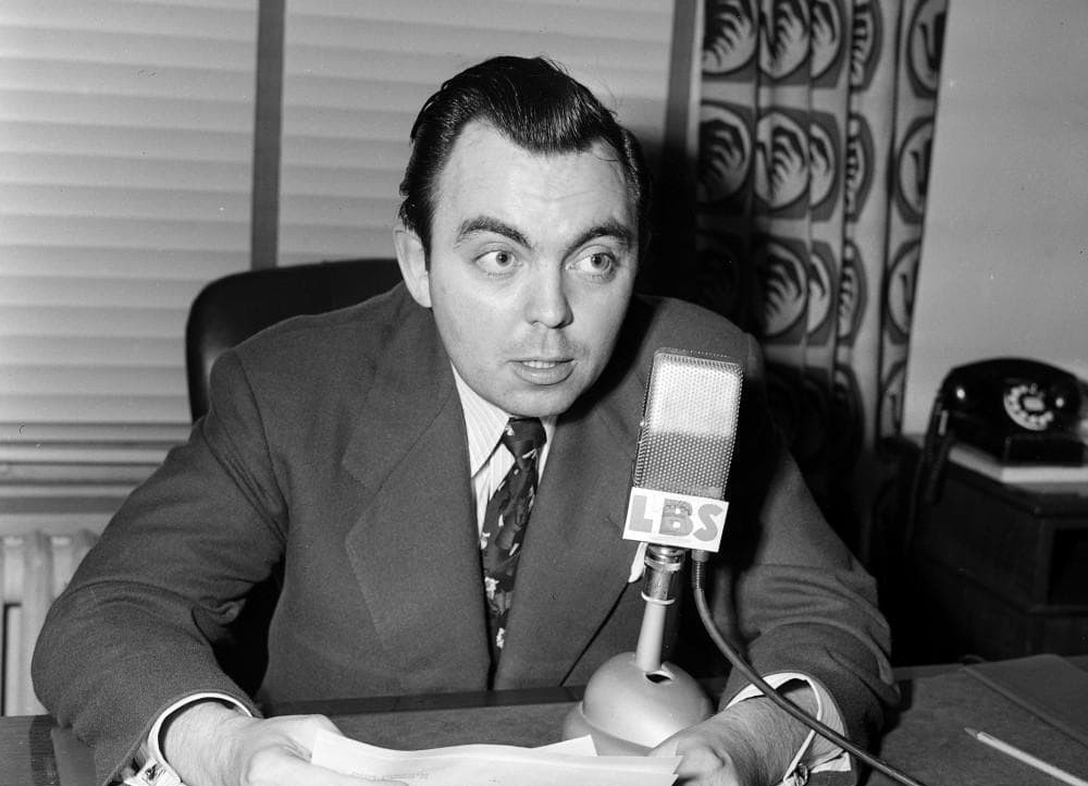A man in a suit speaking into a vintage microphone with 'LBS' labeled on it, seated at a desk with papers and a rotary phone, in a room with patterned curtains.