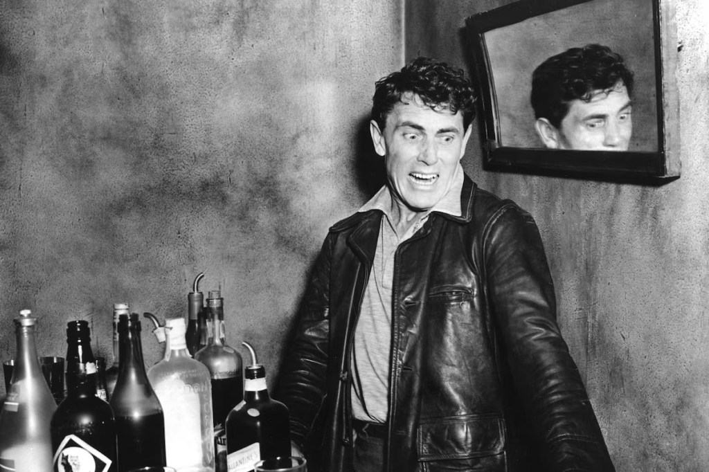 A man in a leather jacket, standing in front of a bar filled with various bottles, appears to be surprised or shocked, with a mirror reflecting his expression in the background.