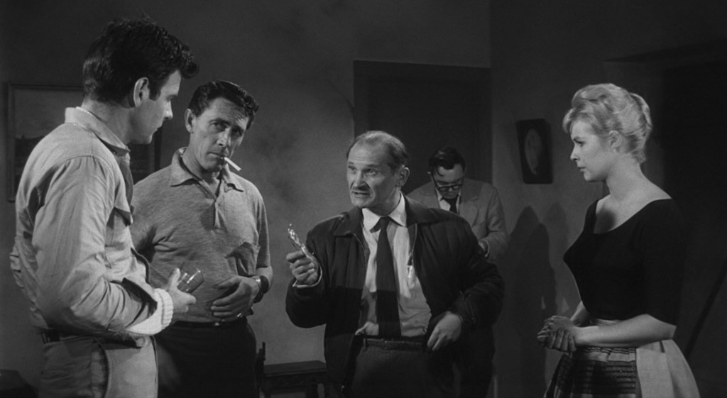 A group of four people in a dimly lit room, engaged in a tense conversation. Two men in casual attire are standing on the left, while a man in a coat gestures with an object in his hand. A woman in a black top stands to the right, observing the discussion.