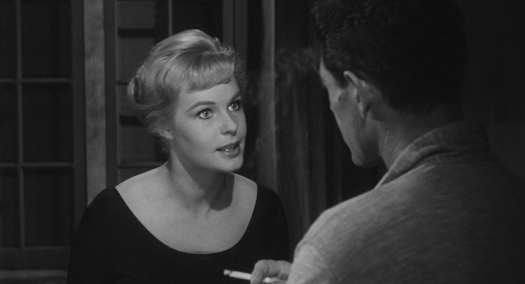 A woman with short blonde hair and a black top is speaking intently to a man who is facing her, in a dimly lit room with wooden features.