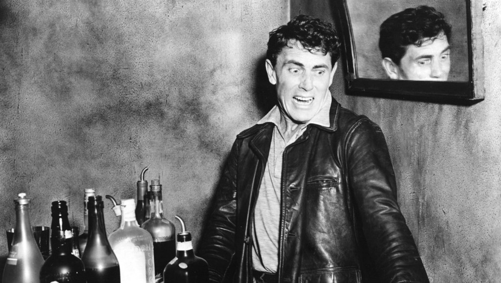 A man in a leather jacket standing in front of a bar with various bottles, showing an exaggerated facial expression with a reflection of his face in a mirror behind him.
