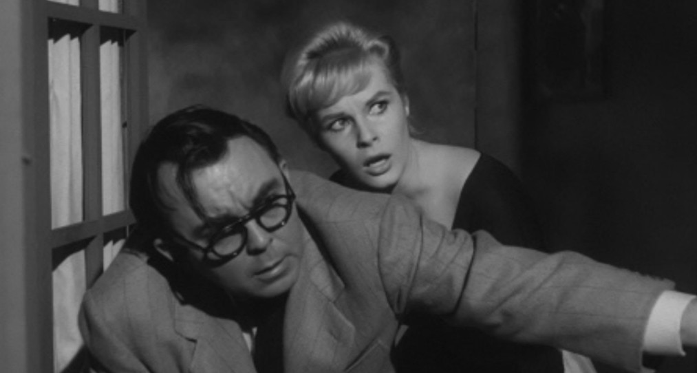 A man in glasses and a suit and a woman in a black dress look anxiously out from behind a window, illustrating a moment of suspense.