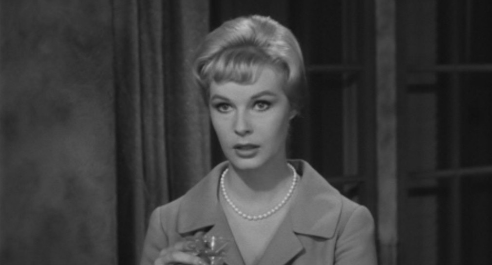 A woman with short, styled hair wearing a pearl necklace and a light-colored coat, looking thoughtfully while holding a drink.