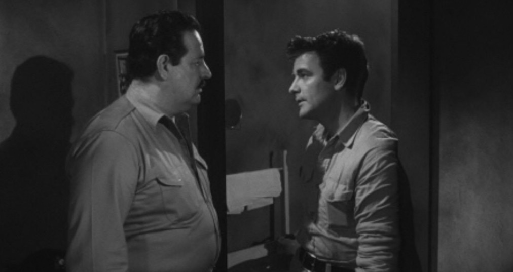 A dramatic black and white scene featuring two men in conversation, one in a uniform and the other in casual clothing, set in a dimly lit room.