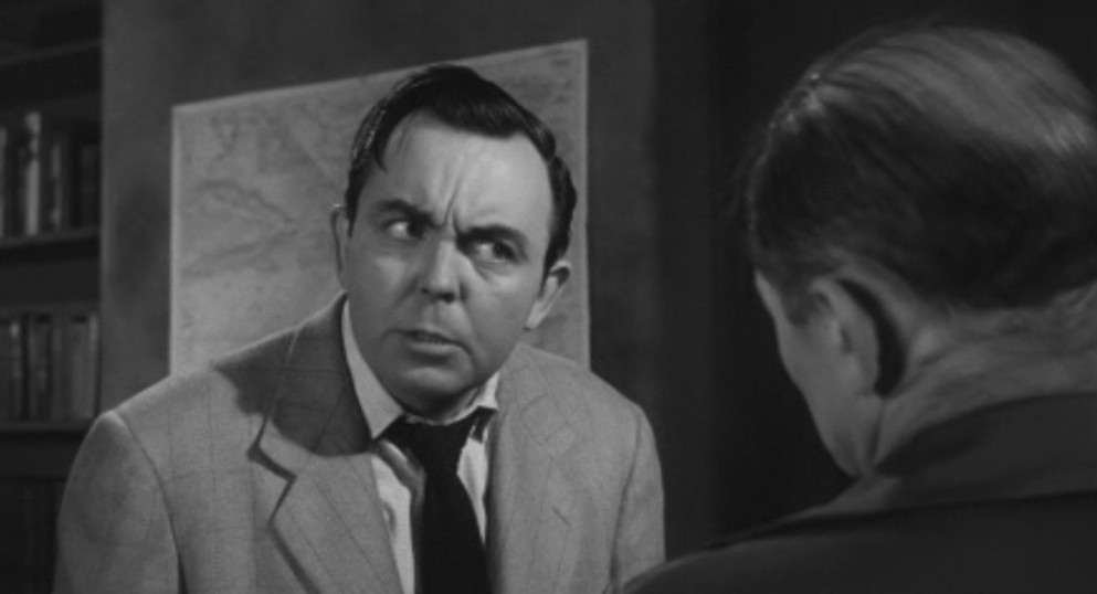 A man in a light suit with a concerned expression engages in a serious conversation with another person whose back is facing the camera. A map is visible in the background.