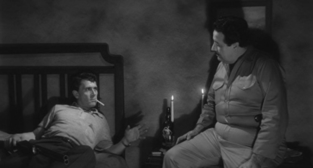 Two men seated in a dimly lit room; one is reclining on a bed with a cigarette, while the other sits beside him, both engaged in an intense conversation.