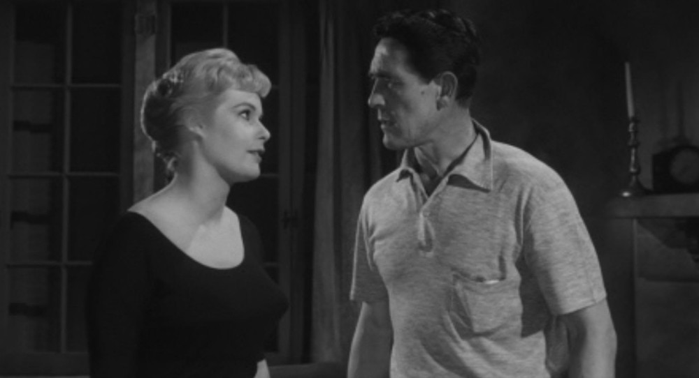 A black and white scene depicting a woman with short, styled hair wearing a dark top, engaged in an intense conversation with a man in a light-colored polo shirt. They are positioned facing each other in a dimly lit room, with a window and shadows in the background.
