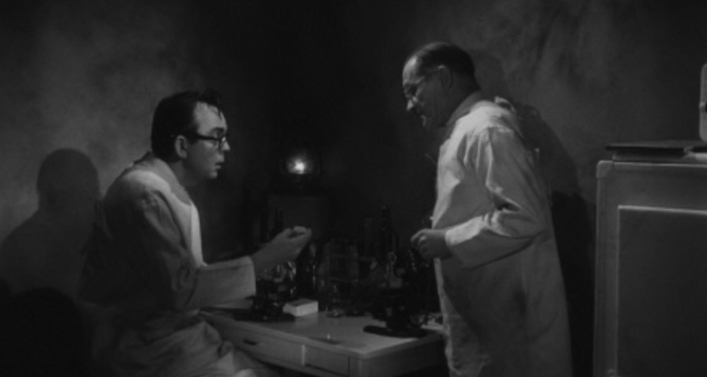 Two scientists in lab coats converse in a dimly lit room filled with lab equipment, one gesturing with a vial.