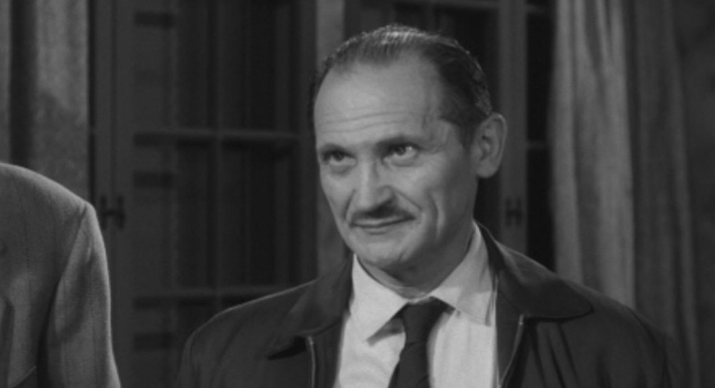 A man in a dark jacket and tie, with a mustache, looking slightly amused or skeptical, in a black and white setting.