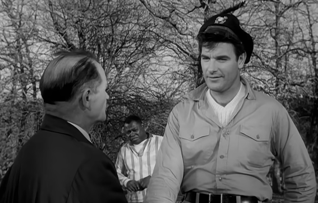 A police officer in a uniform interacts with a man in formal attire, while another man in striped clothing stands in the background among bare trees.