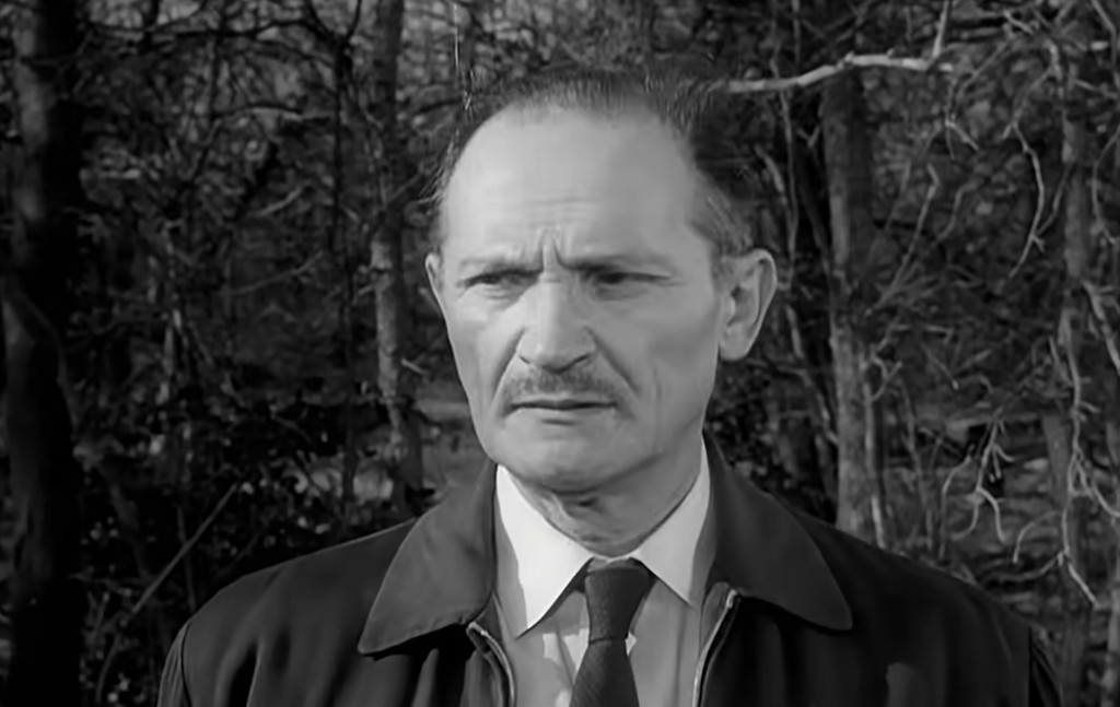 A close-up of a middle-aged man with a serious expression, set against a blurred forest background. He has a mustache and is wearing a suit and a coat.