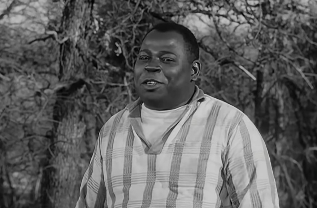 A man wearing a striped shirt stands outdoors, smiling amidst a background of trees.