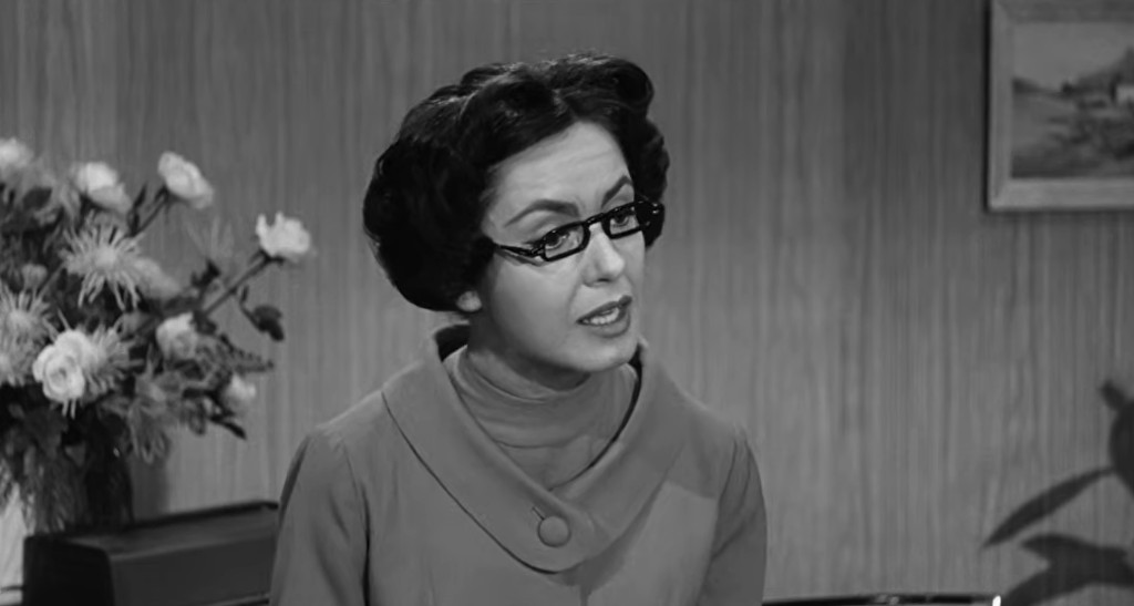 A woman with short, dark hair styled in curls wearing glasses and a grey dress, speaking in an indoor setting with a wooden wall and flowers in the background.