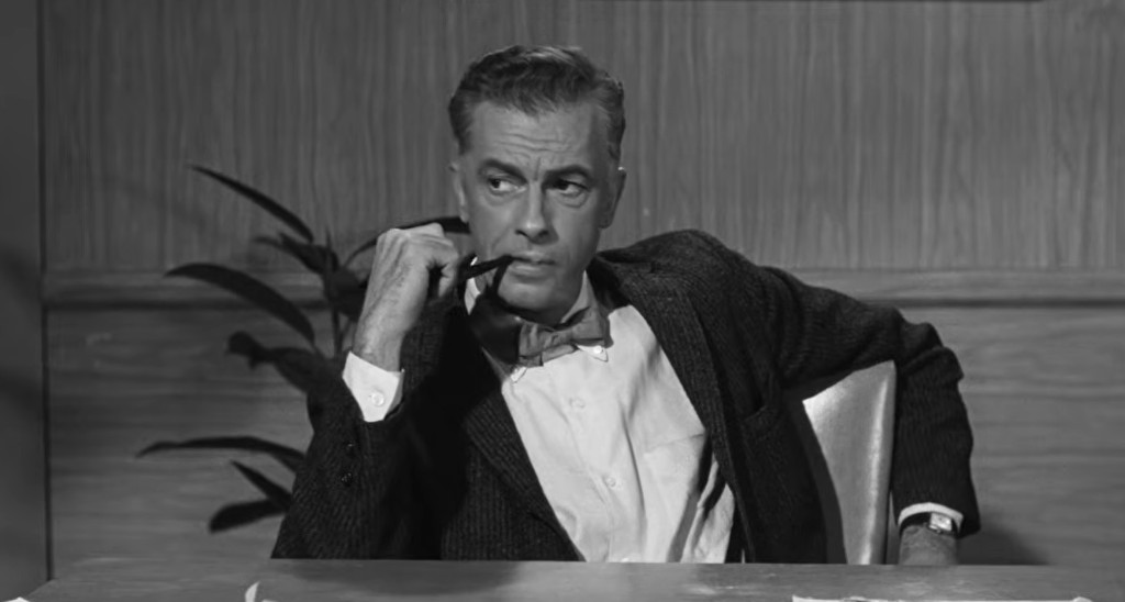 A man in a bow tie and jacket sitting at a table, thoughtfully holding a pen to his chin, with a wall plant in the background.