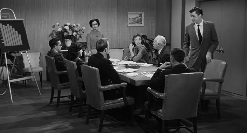 A black and white image of a business meeting around a long conference table, featuring eight people, including men and women, engaged in discussion. A woman stands beside a seated man, looking at a woman across the table while a chart is displayed on an easel in the background.