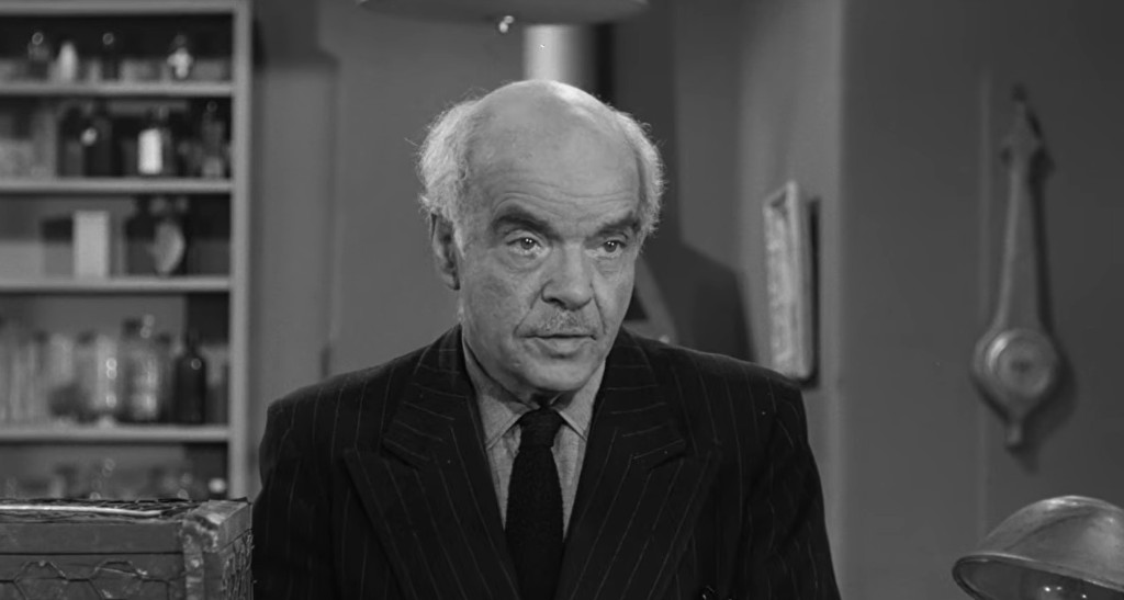 A man with gray hair and a mustache, wearing a black pinstripe suit, appears serious while standing in a room filled with various bottles and objects in the background.