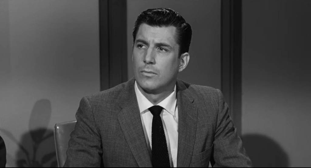 A man in a 1960s suit and tie, showing a serious expression while seated at a table.
