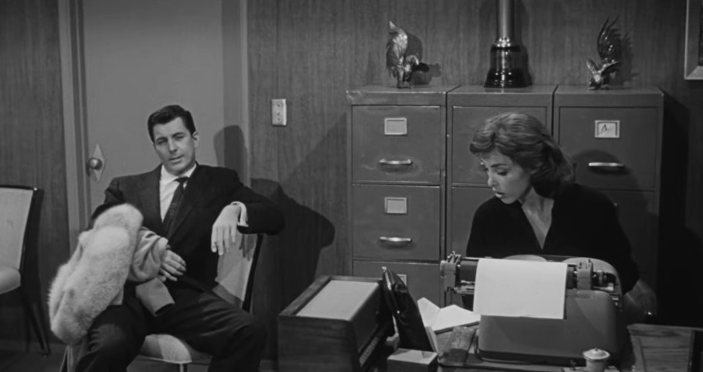 A well-dressed man sitting on a chair with a fur coat draped over his arm, looking pensive, while a woman in a black dress types on a typewriter at a desk in an office setting.
