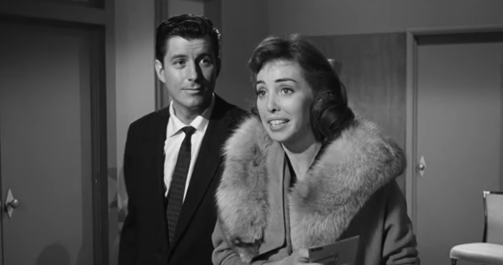 A man in a suit and a woman in a coat with a fur collar are standing in a well-lit interior, appearing to engage in conversation.
