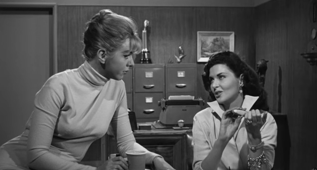 Two women sit in an office, engaged in conversation. The woman on the left, with blonde hair, wears a light-colored turtleneck, while the woman on the right, with dark hair, is dressed in a white blouse and gestures with her hands. The background features office furniture and decor.