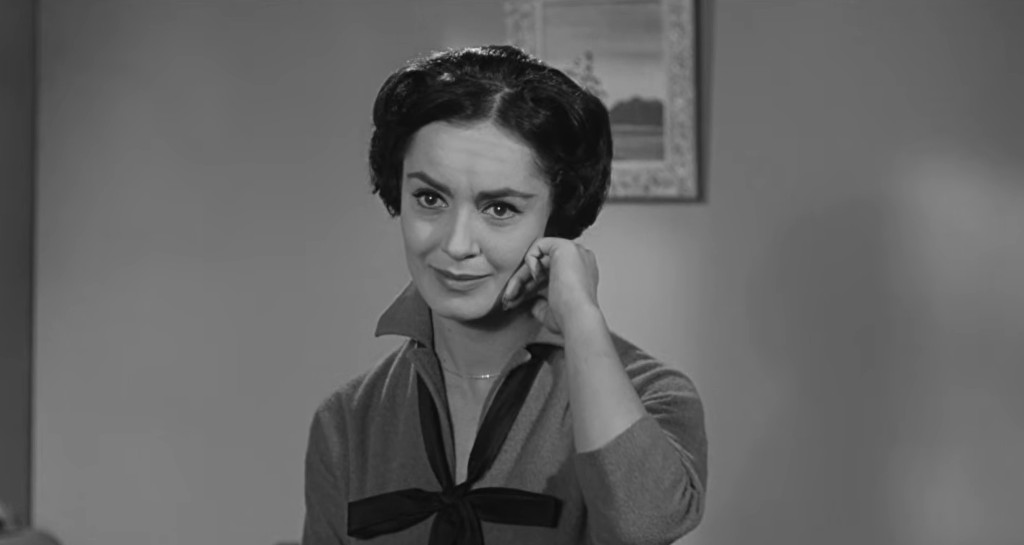 A woman with dark hair styled in soft waves, wearing a gray outfit with a black bow, smiling gently while resting her chin on her hand in a softly lit interior.