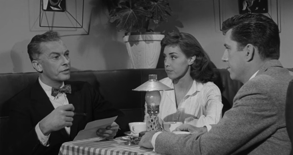 A man in a suit seated at a table with a notepad, speaking to a woman in a white blouse and a man in a gray jacket, in a café setting with a lamp and potted plant in the background.