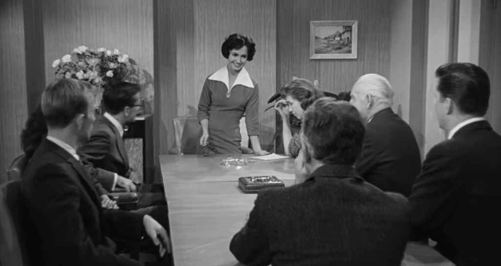 A group of people seated around a conference table in a mid-century modern office. A woman in a red dress with a white collar stands at the head of the table, smiling, while others around her listen attentively.