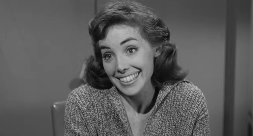 A woman with curly hair smiles cheerfully, wearing a knitted sweater, in a black and white setting.