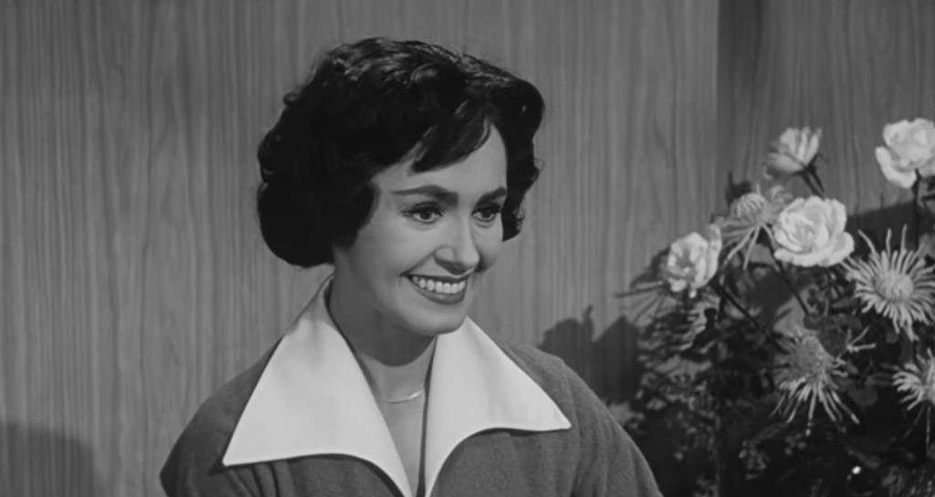 A woman with short, curly dark hair smiles warmly, wearing a shirt with a white collar, in front of a backdrop featuring flowers.