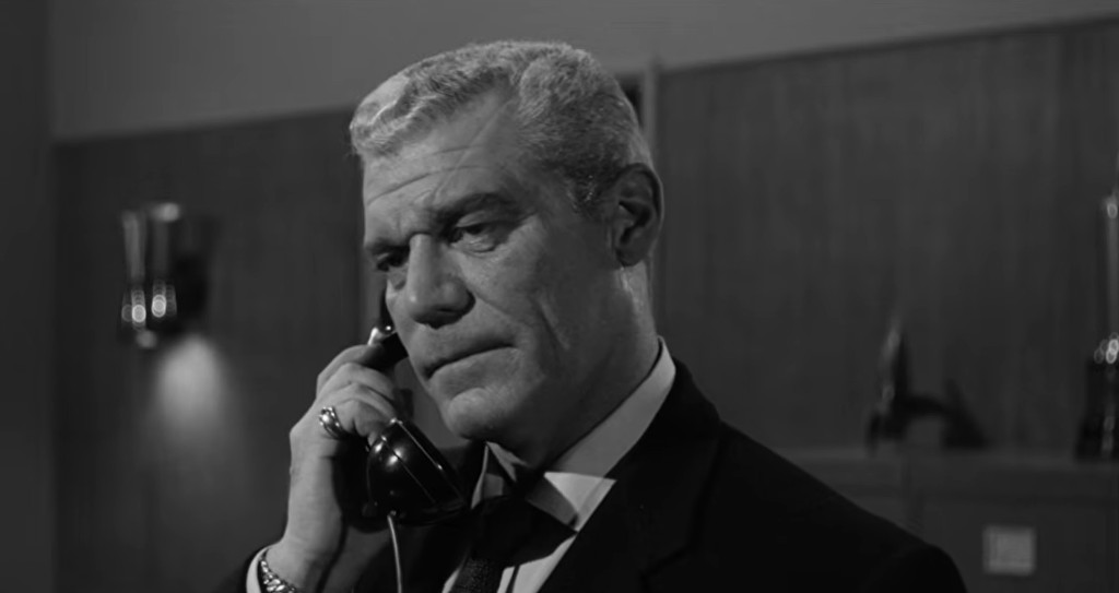 A man in a suit speaking on a rotary telephone, with a serious expression in a dimly lit office.
