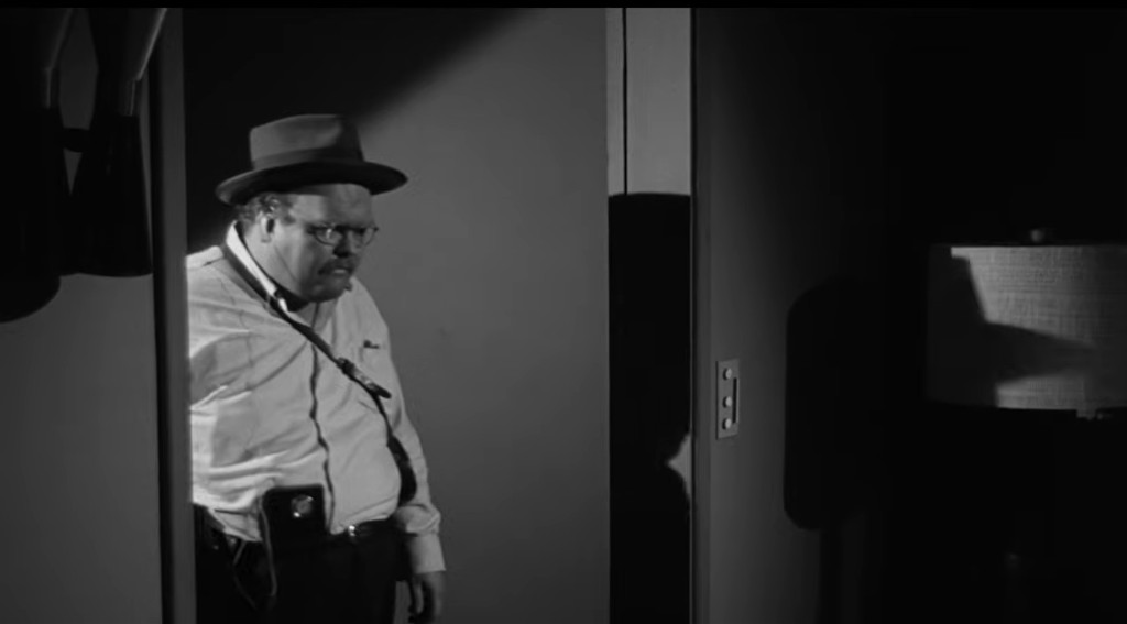A man wearing a hat and glasses stands in a doorway, looking serious, with a shadow cast behind him and a lamp visible in the background.