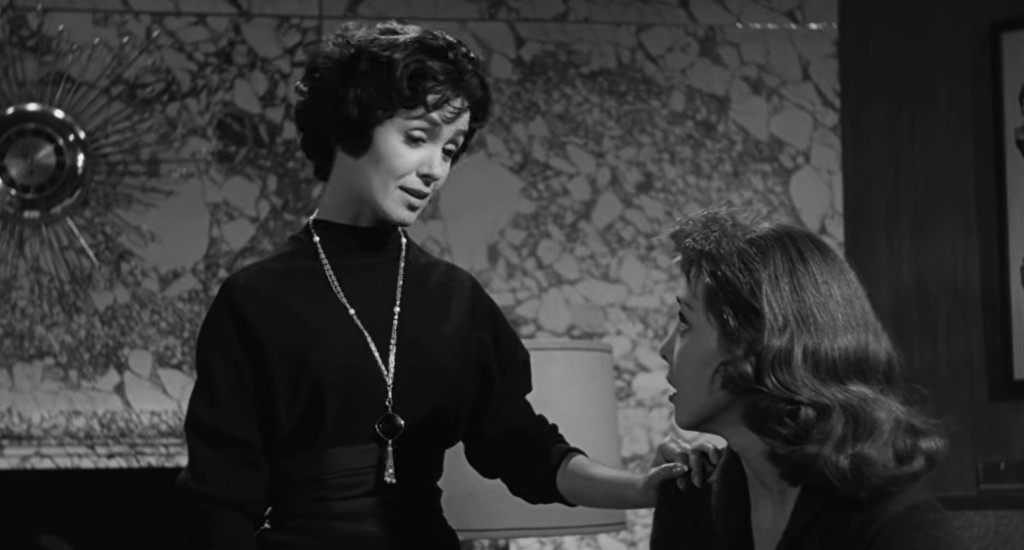 A black and white scene featuring two women engaged in conversation. One woman, dressed in a dark top and necklace, is speaking to another woman with wavy hair, who appears to be listening intently. The background shows a decorative wall with a large sunburst design.