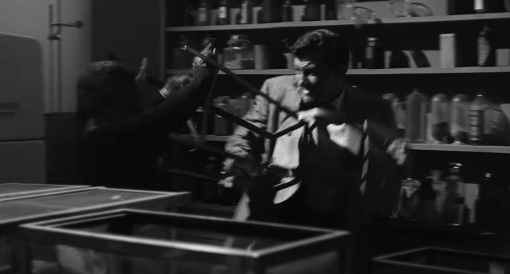 A tense scene in black and white depicting a struggle between two people, one wielding a chair leg as a weapon, set in a cluttered room filled with various bottles and containers.